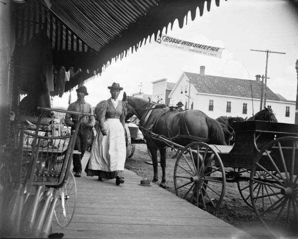 View from sidewalk of a horse-drawn buggy parked along the side of Main Street, looking southeast at First Street. An immigrant couple, possibly Norwegian, are walking up the board sidewalk of town under the awning of a storefront. A baby carriage and other items are displayed along the front of storefront, and clothing hangs from the awning supports. A banner across Main Street advertises the "La Crosse Inter State Fair, Sept. 14, 15, 16, and 17, Admission 25 Cents."