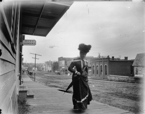 Woman wearing a large flower hat and holding an umbrella is walking on a board sidewalk of Main Street, passing the City Library. The brick office building of Mr. Price is reportedly on the left, on the south west corner of the intersection of Main Street and Second Street. Further down from the library (by the black patch behind the telephone) was Iver Erickson's shoemaker shop. He was an expert and made the caulked log driver shoes.