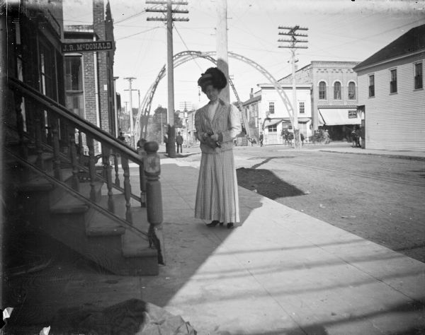 Woman posing standing on the west side of South First Street in front of the photographic studio of Charles J. Van Schaick. There is a sign on the building for J.R. McDonald, and an arch over the intersection of Main Street and First Street.