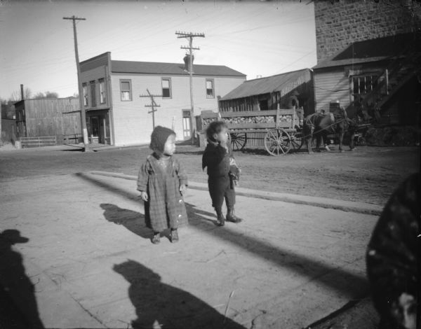 View of two small Native American children standing on the city scale. Across the street is a horse-drawn wagon near commercial buildings.