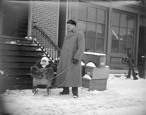 Man and small girl in a homemade sled in front of the steps that lead to the photographic studio of Charles J. Van Schaick. Storefront on the left sells cream separators. Man identified as probably Frank Parsons, the American Express agent, with his daughter.