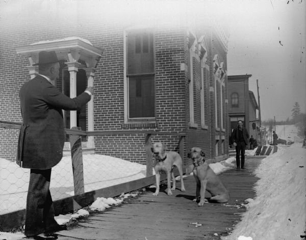 Two men playing with two dogs on sidewalk on the west side of South First Street between drifts of snow. Hardware store partially obscured on the northwest corner of the intersection of Main and First Streets.