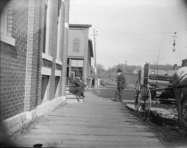 View of the north and south corners of the west side of the intersection between First Street and Main Street. Jones and Murray Hardware store identified on the northwest corner. The man with the long gray beard on the right is identified as Charles Felt.