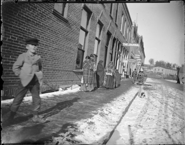 A boy is standing on a snow-covered wooden sidewalk on the left, and Native American women wrapped in blankets are walking on the sidewalk on the right. View of North Water Street from Main Street, before 1911. Brick building includes offices for The Journal, and the wooden building next to it holds the store of M.F. Stebbins, milliner. In the distance is a sign for the Freeman House.