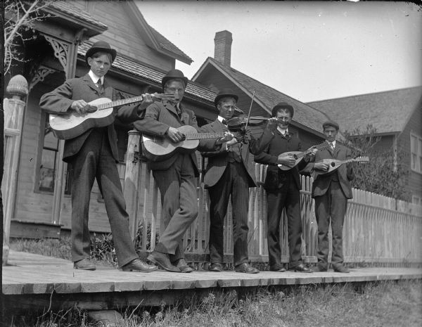 Five men playing string instruments while standing on a town sidewalk. They are identified from left to right as: unidentified man, Anton Hagen, Ole Strand, John Strand, and Albert Hanson. They are standing side by side, in dress clothes with bowler hats, on a raised boardwalk with weeds beneath, and two houses behind them.