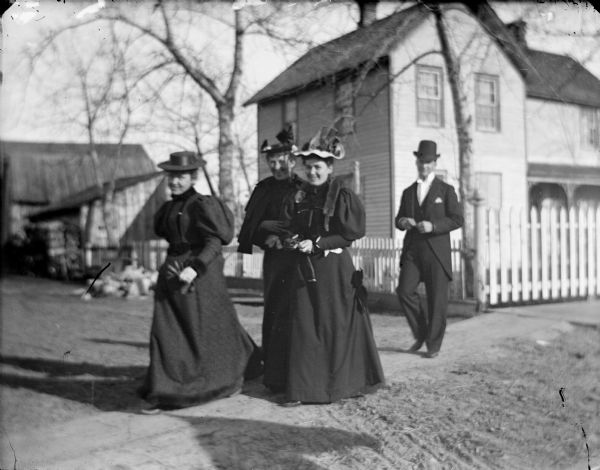 Three women and a man crossing a dirt street.