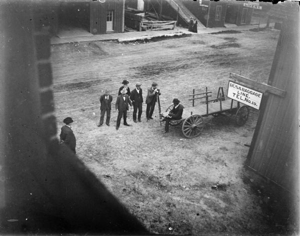Elevated view from the Van Schaick's studio of seven men with a surveying instrument, probably on South First Street. Two women are walking on the wooden sidewalk across the street. Signs for business include the Bus and Baggage Line at Tel. No. 13, and the Waterson (?) Livery.