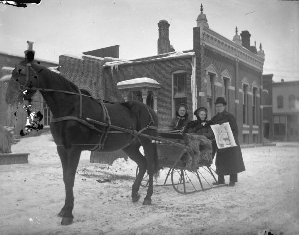 Two women in a horse-drawn cutter sleigh on a snowy street. A man is posing standing nest to them, displaying a Charter Oak Mills advertisement/calendar.