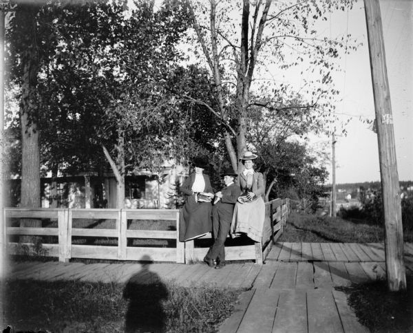 One man and two women holding books and sitting on a fence at the corner of Fourth Street and Harrison Street.