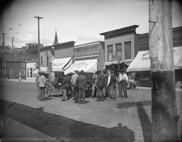 Crowd of men examining an automobile on Main Street, possibly the vehicle that belonged to Dr. Krohn. Storefronts identified, from left to right, include: a barbershop, City Bakery, Kelly's, Sign for Werner Drugstore, The Fair, Confectionery, Mo(?) Groceries.
