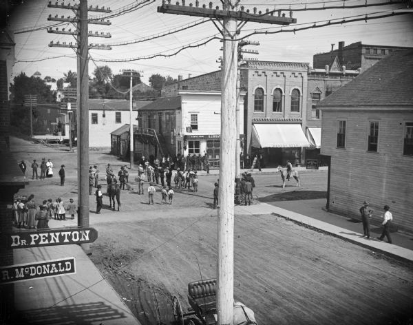 Crowd gathered at the intersection of Main Street and First Street for a performance by the town band, around a man holding a dog wearing a coat with words on it. Signs in the lower left corner are for Dr. Penton and R. McDonald. Storefronts on Main Street, from left to right, include the O.J. Heath Sample Room, Moses Paquette Store, and a clothing store. A.P. Johnson store located on the southeast corner of the intersection.
