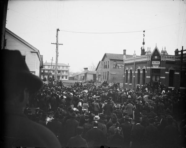 Large crowd milling around a band comprised of African Americans at the intersection of First Street and Main Street, and showing South First Street. Identified businesses from left to right include: a livery, the Merchants Inn at the end of First Street, the Charles J. Van Schaick Photography Gallery with a sign for Dr. Krohn, and the Jackson County Bank.