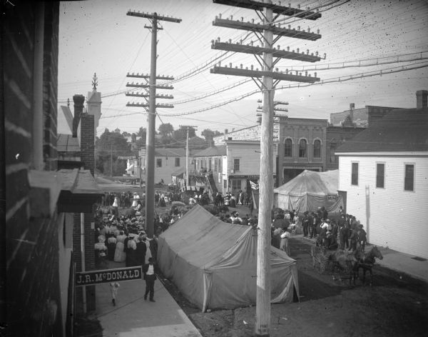 Crowd gathering around tents on South First Street and Main Streets for a special occasion. Sign in lower left for J.R. McDonald and the O.J. Heath Sample Room on the northeast corner of the intersection of Main Street and First Street.
