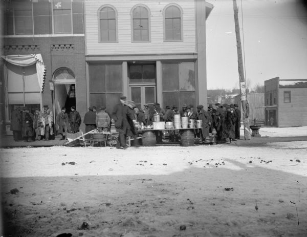 Men gathered around tables covered with pots and pans on the snow-covered street in front of a hardware store.