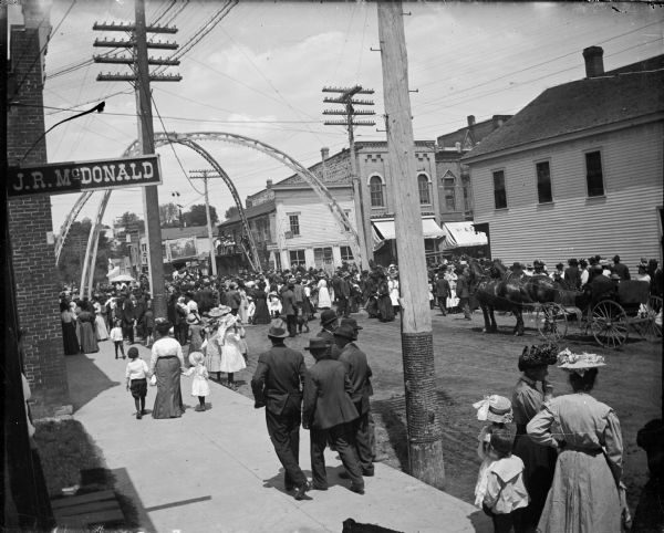 Crowd watching a circus performance in the street, possibly centered on a gorilla named Johanna according to the banner in the background. There is an arch over the intersection. In the foreground on the left is a sign for J.R. McDonald. Storefronts on Main Street identified from left to right include: a Saloon, Mose Paquette Store, and A.F. Werner.