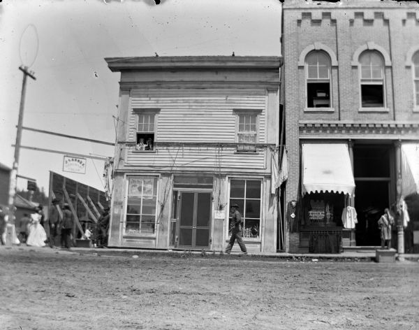 Storefronts identified from left to right include: J. Sannes Merchant Tailor with a sign on building for "Lemonade and Pop," and an unidentified storefront with a cigar store Indian in front of it.