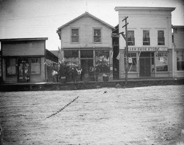 Storefronts identified from left to right include: unidentified, Thomas Hendricks' Barbershop, and the Cash Shoe Store of Locken and Giere. All the storefronts are decorated for a patriotic celebration with United States flags. An orange tree and lemon tree are in large wooden pots in front of the barbershop.
