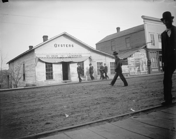 Men walking in front of Squire's Oyster House restaurant, east on Water Street. Posters advertise the "Georgia Up-To-Date Minstrels" at the Opera House on Saturday, April 7.
