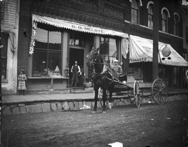 Horse and wagon in front of a storefront on Main Street with an elevated board sidewalk. Storefronts identified from left to right as G.G. Melbye Groceries and J.A. Eckern Jeweler.