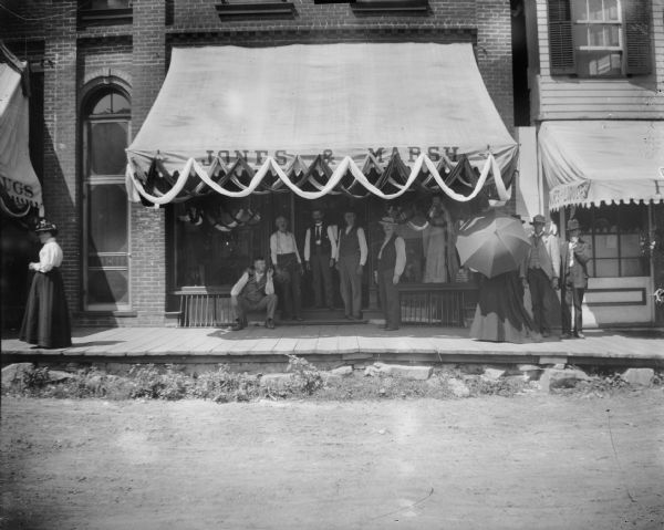 View from street towards a group of people poing in front of the storefront of the Jones and Marsh Dry Goods. John Marsh, the proprietor, is sitting on the left.