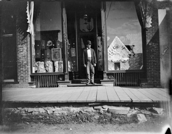 View from street towards a young man wearing suit and a derby hat posing standing in the doorway of Jones and Marsh Dry Goods.
