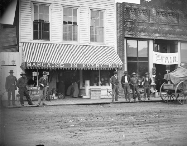 Men posing in front of stores. Businesses from left to right are the A.F. Werner Drugstore and Clothiers, and the Fair Store. The former has the slogan "Bargains In Everything" displayed on its awning.
