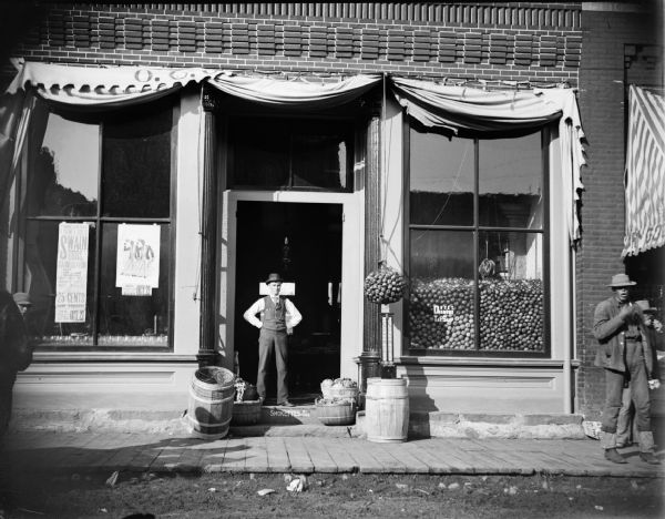 O.C. O'Hearn Grocery Store, Main Street, with the front window filled with apples, and a man standing in the doorway. Another man is standing on the sidewalk on the far right. Posters in the window advertise: "Swain Brothers Carnival of Fun on Tuesday, October 23."