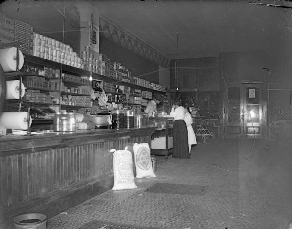 Interior of a dry goods store, possibly Monsos Brothers. Woman and girl purchasing supplies.
