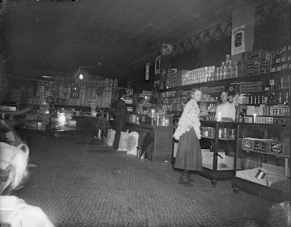 Interior of a dry goods store, possibly Monsos Brothers. Woman and man purchasing supplies. Man behind the counter identified as probably Martin Olson.