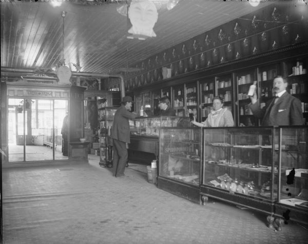 Man purchasing medicine in a drugstore. Two men and one woman behind the counter, all holding up products. Individuals identified, from left to right, as probably two unidentified men, Mrs. Roddy, and Ben Warner.