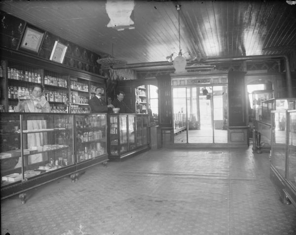 Interior of a drugstore. Two men and one woman behind the counter. Persons identified, from left to right, Mrs. Roddy, Ben Warner, and an unidentified man.
