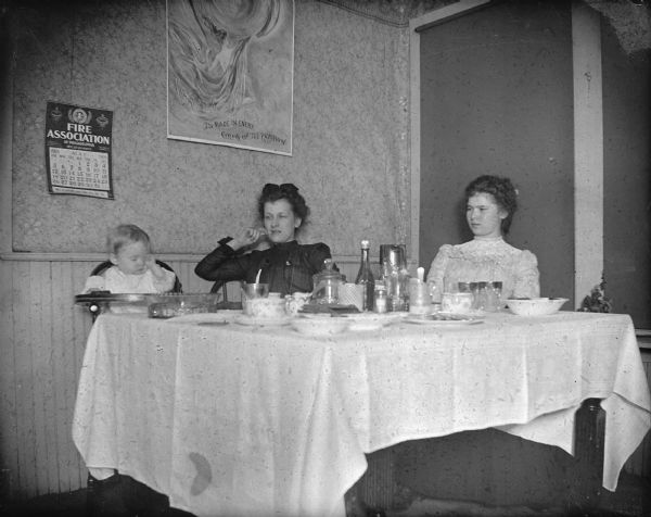 Two women and a child in a highchair sitting at a table, possibly in a restaurant. Woman on the left is using a toothpick. Calendar on the wall from the Fire Association of Philadelphia, showing the month of May, 1901, and poster, possesses a quote, "Is Made in Every Color of the Rainbow."