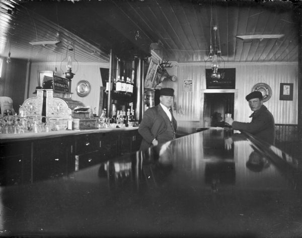 Man having a drink at a bar. Location identified as a tavern that later became the Cozy Corner. Calendar on the wall shows the month of January. Man on left identified as Andrew Johnson.
