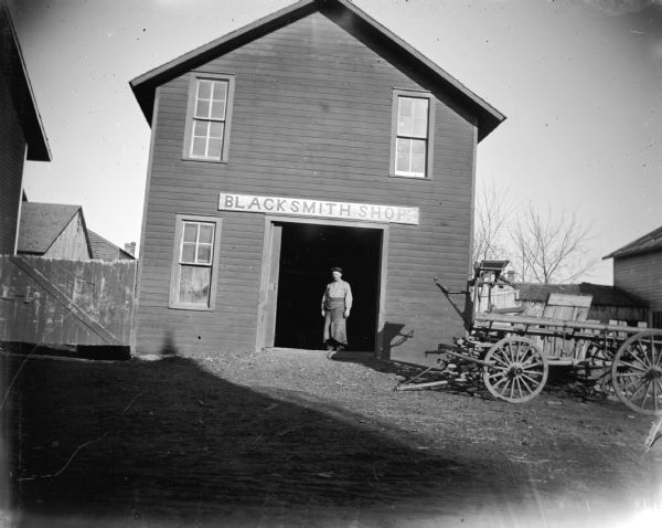 Blacksmith in apron standing in the doorway of a blacksmith shop. Man identified as possibly Fred Weller.