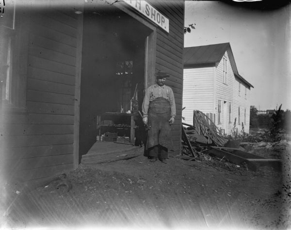 Blacksmith in apron standing just outside the doorway of a blacksmith shop. Man identified as possibly Fred Weller.