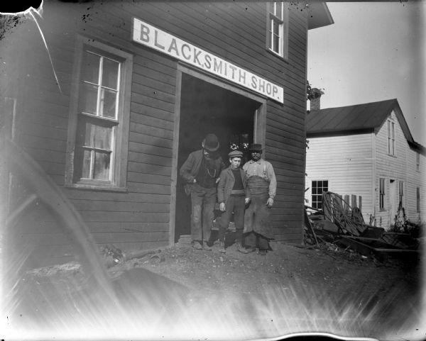 Man wearing a derby, a boy, and a blacksmith wearing an apron, standing just outside the doorway of a blacksmith shop. Blacksmith identified as probably Fred Weller.