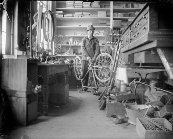 Man repairing a bicycle in a shop, probably the Repair Shop of Leslie Werner. Man identified as probably Leslie Werner.