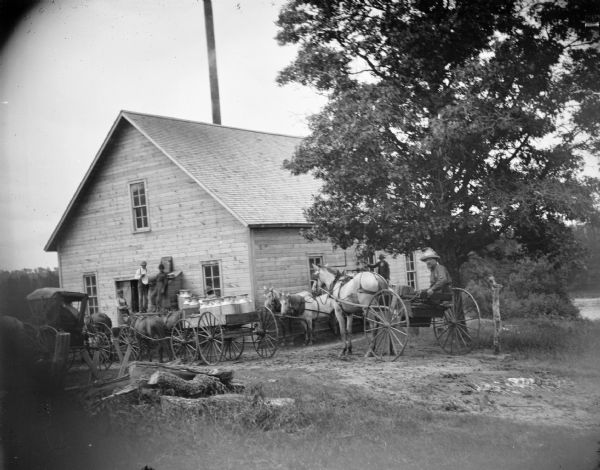 Men bringing cans of milk to a building, probably a creamery by a river.