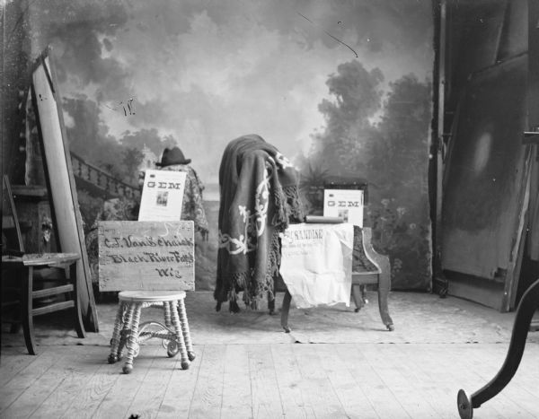 Studio portrait of signs that advertise "GEM," list "C.J. Van Schaick, Black River Falls, Wis.," and a hat. GEM may be a manufacturer of photographic equipment. There is a painted backdrop in the background.