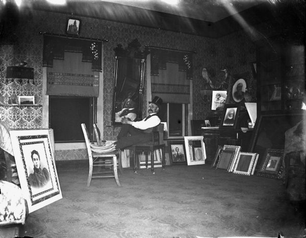 Man in top hat sitting in front of mirror, surrounded by portraitures taken inside the photograph studio of Charles J. Van Schaick. Possible photograph of the Jackson County Court House in the upper left. Man identified as probably Charles J. Van Schaick of Thomas D. Van Schaick.