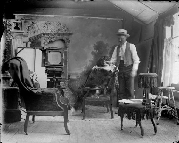 Man petting dog, who is sitting on a chair, inside the photographic studio. There is a large studio camera on the left, several pieces of studio furniture, a painted backdrop, and large windows on the right. Man identified as possibly Olaf Olson.