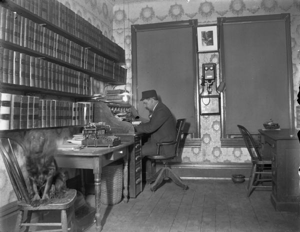 A man, possibly John Forbes, working at a roll-top desk in his office over the old Fire Hall. A dog is sitting on a chair in the lower left. Books on the shelves include American Digest 1900. There is a typewriter on the table, and a telephone on the wall.