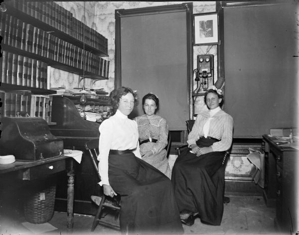 Three women seated in front of a roll-top office desk, possibly the office of John Forbes over the old Fire Hall. A covered typewriter on the table and a telephone on the wall.