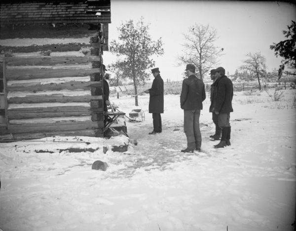 One man armed with a shotgun, facing a man in the doorway of a log house, and surrounded by three other men standing in the snow.