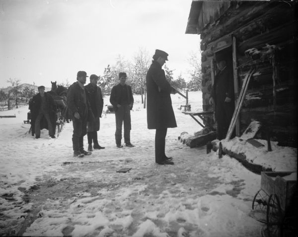One man armed with a shotgun, facing a man in the doorway of a log house, and surrounded by four other men standing in the snow, also a horse and sleigh.