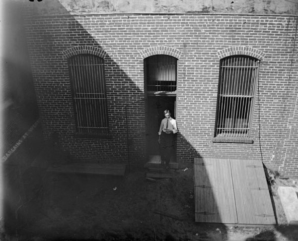 Man standing in the open rear barred doorway of the Jackson County Bank, holding a piece of watermelon, surrounded by barred windows that have panes and curtains. Entrance to the building cellar is on the right, foreground. In the left center is a sign proclaiming "Bargains at Werner."