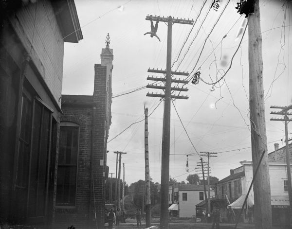 Man hanging upside down from the top of a telephone pole with spectators below. O.J. Heath Saloon on the northeast corner of the intersection of First and Main Streets.