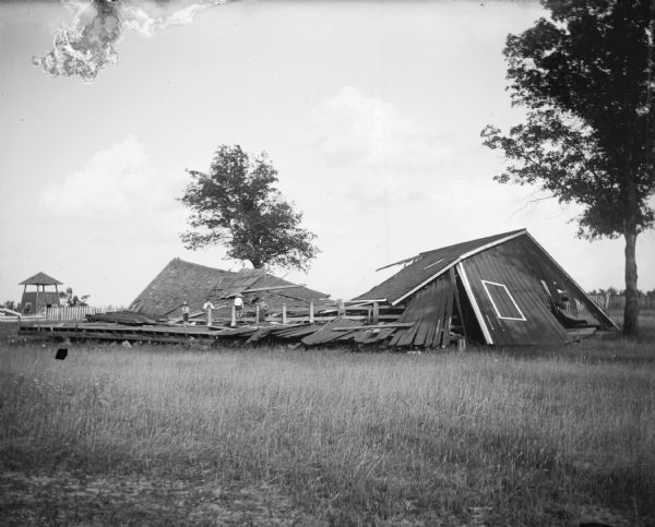 Four boys are posing standing and sitting among the debris of a collapsed wooden building at the Jackson County Fairgrounds, probably caused by a tornado. There is a pavilion in the background on the left.