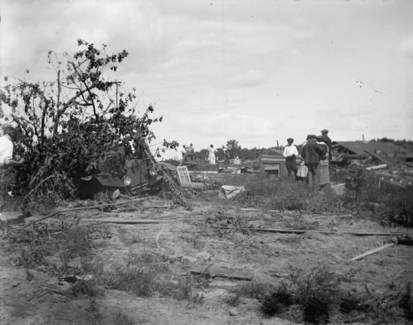 Group of men and women looking at the debris of a building, possibly a house, destroyed by a tornado. Wreckage of an automobile on the left.