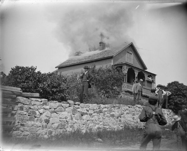 Frame house on fire, and men standing on stone fence in front of the burning building.